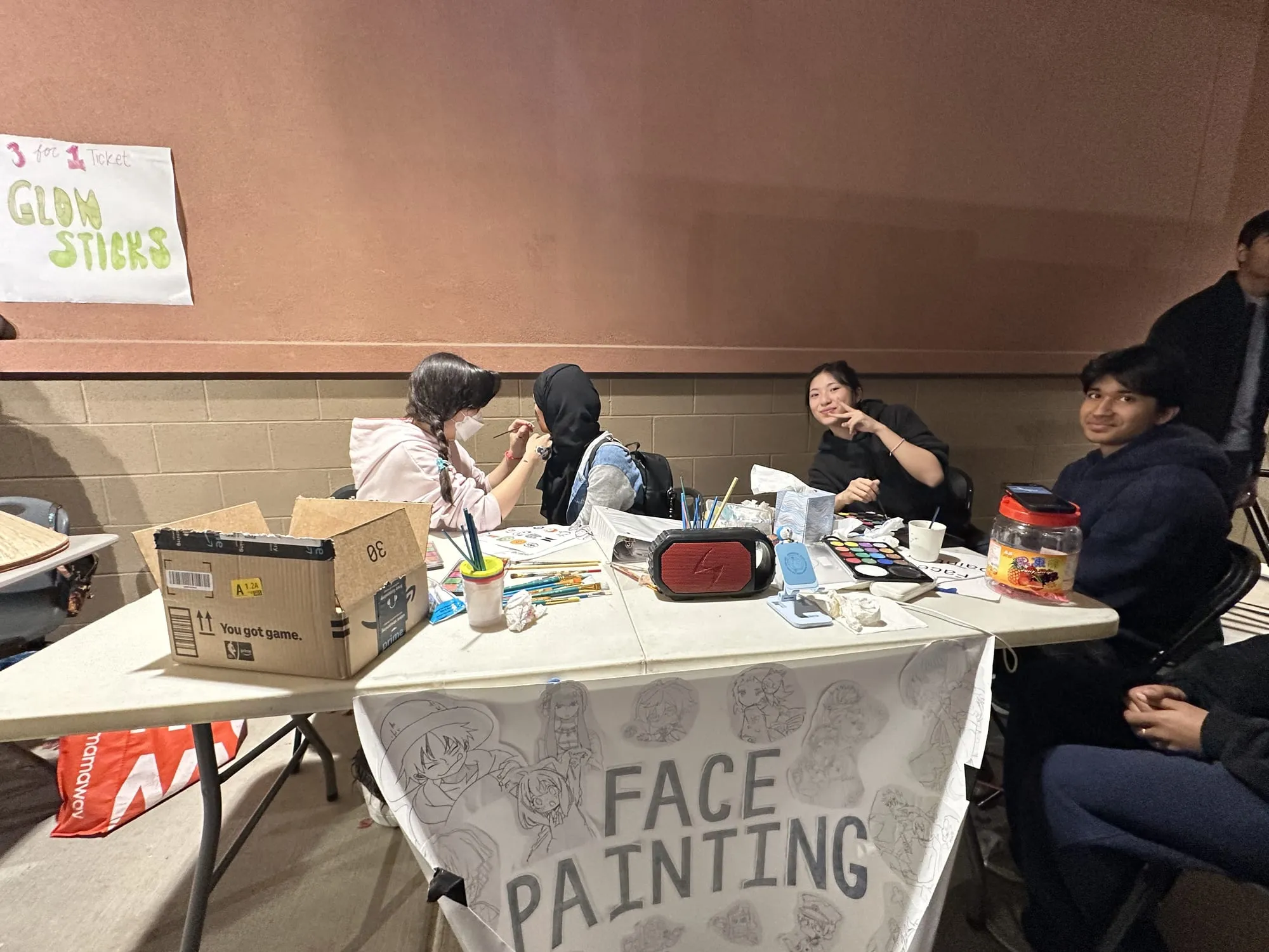 Students paint faces at a table with a hand-lettered face painting banner