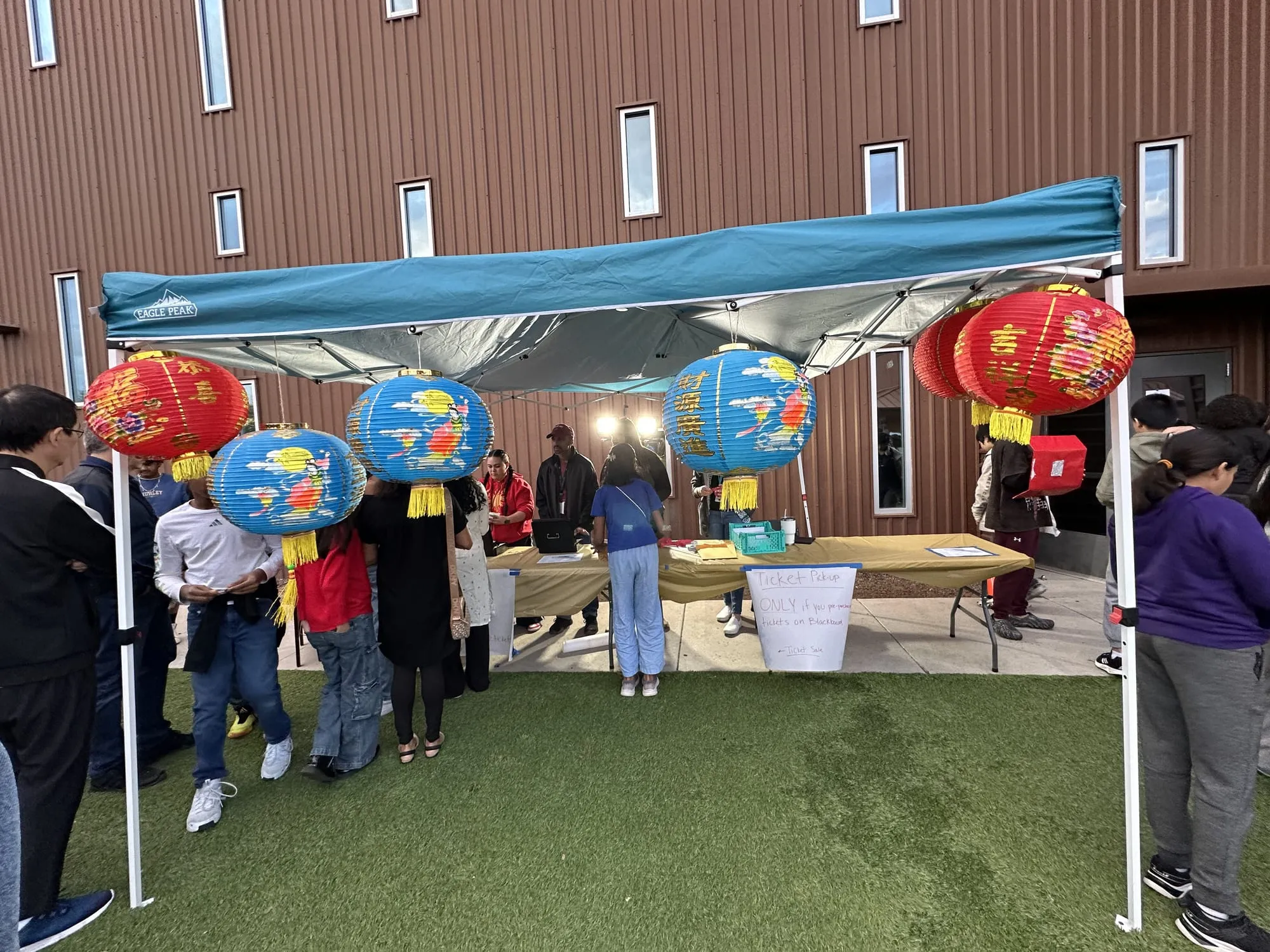 A booth under a canopy hung with red and blue paper lanterns, with a line of visitors