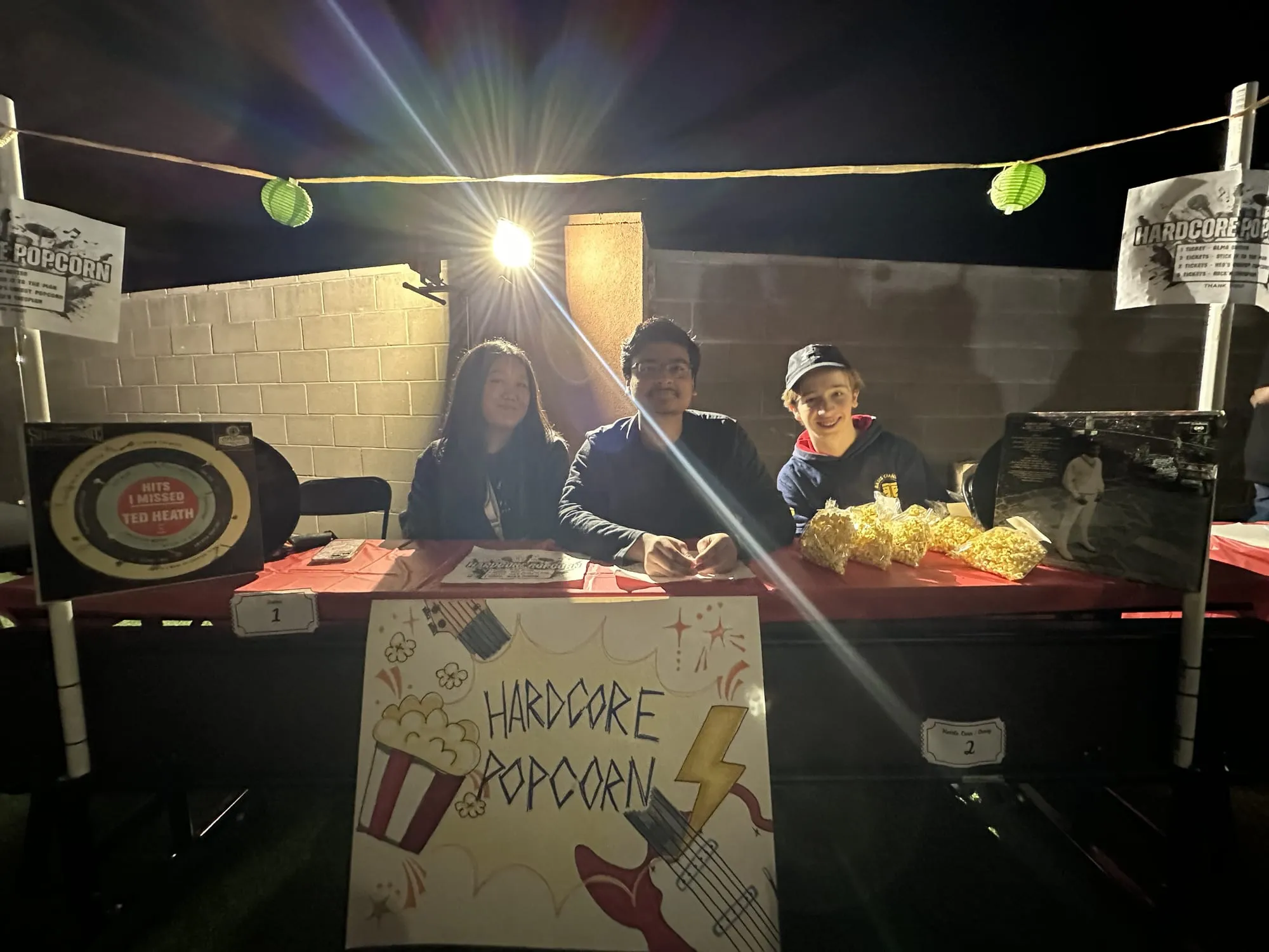 Three students at a popcorn booth lit by string lights at night